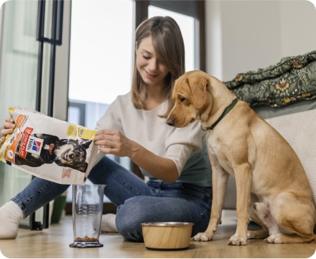 O femeie toarnă hrană Hill's Pet Nutrition într-un recipient de măsurare, în timp ce un câine o privește cu atenție.