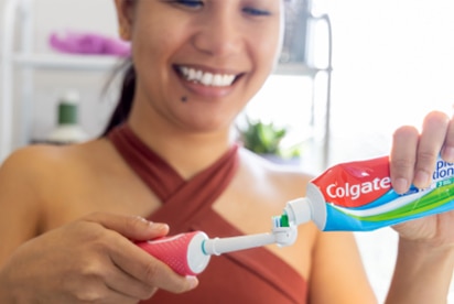 O femeie zâmbind în timp ce pune pastă de dinți Colgate pe o periuță de dinți într-o baie.